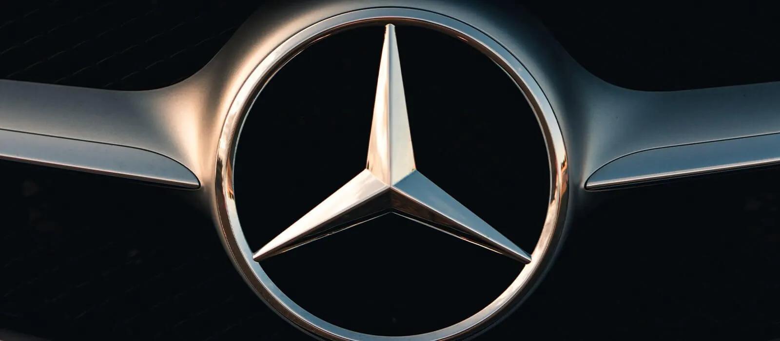 Nahaufnahme eines Mercedes-Benz Logos vor dunklem Hintergrund.